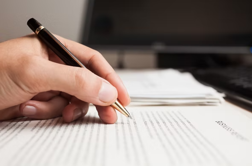 A close-up shot of a person's hand holding a pen over a document. The hand is poised as if writing or signing something on a paper with lines of text visible. A laptop or computer screen is blurred in the background.