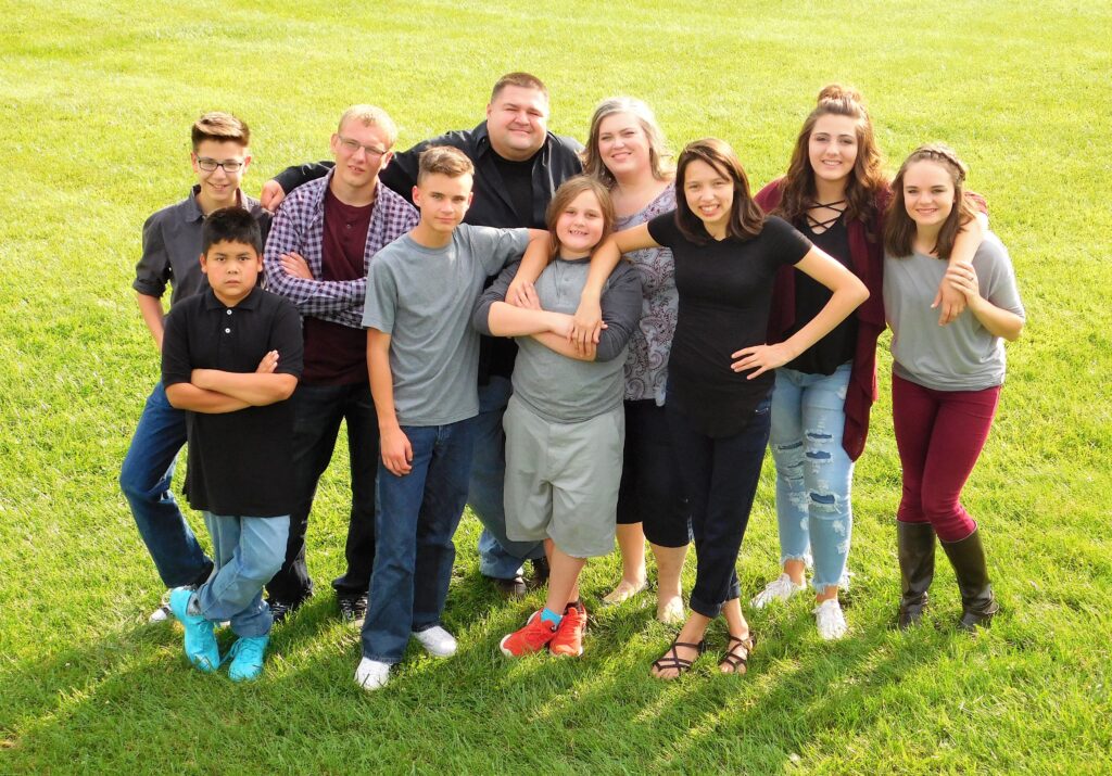 A family of ten people smiling and standing in a grassy field on a sunny day. The group includes several adults and children, all wearing casual clothes.
