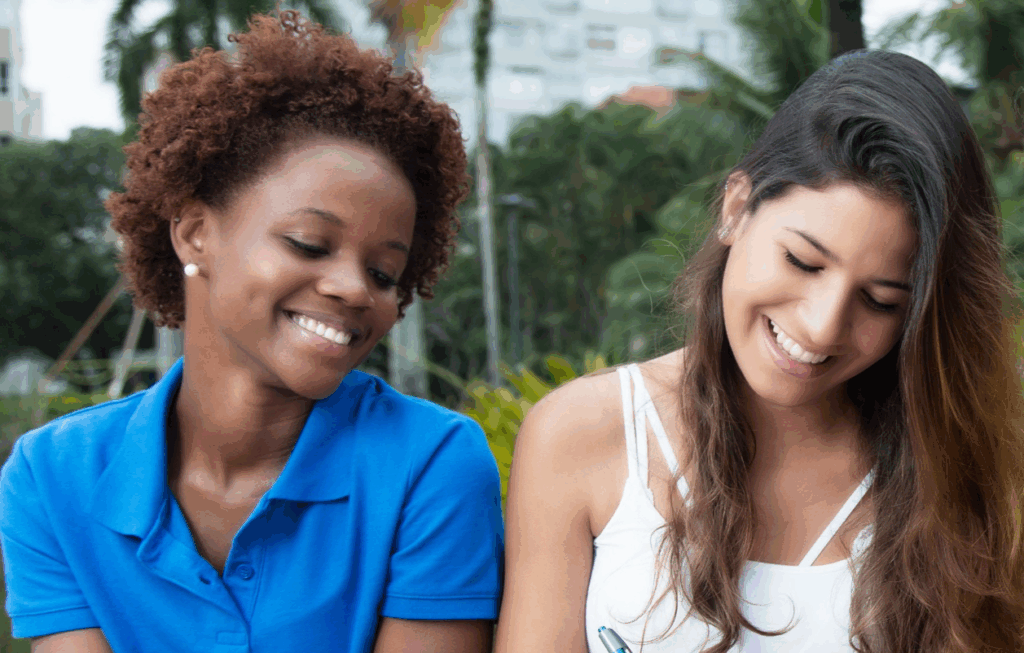 Two young women, one with brown curly hair and the other with straight brown hair, smiling and looking down at something together. They appear to be studying or looking at a document.