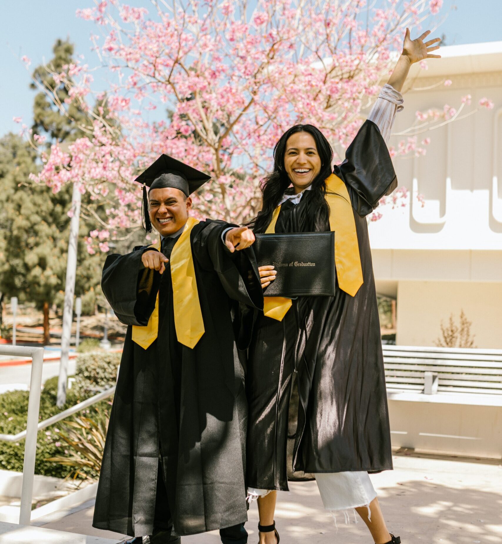 Two happy college graduates in a cap and gown celebrating with a diploma in front of a blossoming tree.